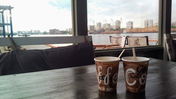 Two disposable coffee cups with wooden stirrers sit on a dark wooden table, with a large window view of an industrial waterside landscape featuring cranes and distant buildings. The setting suggests a relaxed cafe environment contrasting with the busy scene outside.