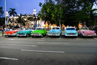 A row of colorful vintage cars parked in an urban area during twilight, with palm trees and an illuminated historic building in the background.