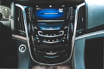 View of a modern car's central dashboard with a glossy black surface. The display screen shows a radio interface, surrounded by various control buttons for audio and climate control. Silver trim lines the edges, and there is a push-to-start button on the left.