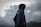 A person wearing a dark rain poncho stands with their back facing the camera against a backdrop of moody, overcast skies and distant mountains. The poncho appears to be made of a water-resistant material and features a circular logo or design. The clouds are thick and gray, indicating a possible impending storm.