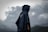 A person wearing a dark rain poncho stands with their back facing the camera against a backdrop of moody, overcast skies and distant mountains. The poncho appears to be made of a water-resistant material and features a circular logo or design. The clouds are thick and gray, indicating a possible impending storm.