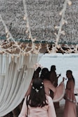 A group of people gather under a thatched canopy by a waterfront, with soft lighting and draped curtains creating a serene atmosphere. The individuals are dressed in matching light pink garments, adorned with delicate floral accents. Twinkling string lights hang above, adding to the festive and elegant setting.
