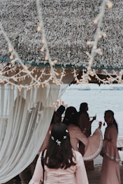 A group of people gather under a thatched canopy by a waterfront, with soft lighting and draped curtains creating a serene atmosphere. The individuals are dressed in matching light pink garments, adorned with delicate floral accents. Twinkling string lights hang above, adding to the festive and elegant setting.