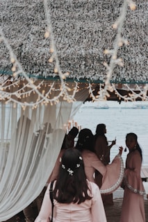 A group of people gather under a thatched canopy by a waterfront, with soft lighting and draped curtains creating a serene atmosphere. The individuals are dressed in matching light pink garments, adorned with delicate floral accents. Twinkling string lights hang above, adding to the festive and elegant setting.