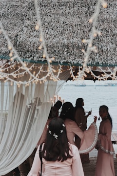A group of people gather under a thatched canopy by a waterfront, with soft lighting and draped curtains creating a serene atmosphere. The individuals are dressed in matching light pink garments, adorned with delicate floral accents. Twinkling string lights hang above, adding to the festive and elegant setting.