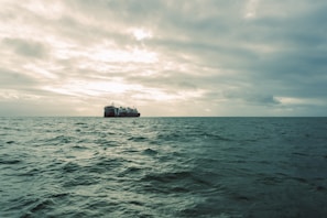 A sleek cargo ship cutting through ocean waves with deep navy sea under a cloudy sky.
