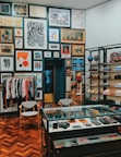 A stylish boutique interior with a well-organized display of clothing, accessories, and art. The walls are adorned with an eclectic mix of framed artworks, creating a gallery-like atmosphere. A rack of clothing is neatly arranged to the left, while shelves on the right showcase folded apparel and shoes. A glass display case in the center contains various small items and accessories. The wooden parquet flooring adds warmth to the contemporary design.