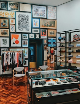 A stylish boutique interior with a well-organized display of clothing, accessories, and art. The walls are adorned with an eclectic mix of framed artworks, creating a gallery-like atmosphere. A rack of clothing is neatly arranged to the left, while shelves on the right showcase folded apparel and shoes. A glass display case in the center contains various small items and accessories. The wooden parquet flooring adds warmth to the contemporary design.