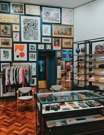 A stylish boutique interior with a well-organized display of clothing, accessories, and art. The walls are adorned with an eclectic mix of framed artworks, creating a gallery-like atmosphere. A rack of clothing is neatly arranged to the left, while shelves on the right showcase folded apparel and shoes. A glass display case in the center contains various small items and accessories. The wooden parquet flooring adds warmth to the contemporary design.