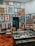 A stylish boutique interior with a well-organized display of clothing, accessories, and art. The walls are adorned with an eclectic mix of framed artworks, creating a gallery-like atmosphere. A rack of clothing is neatly arranged to the left, while shelves on the right showcase folded apparel and shoes. A glass display case in the center contains various small items and accessories. The wooden parquet flooring adds warmth to the contemporary design.