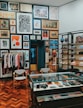 A stylish boutique interior with a well-organized display of clothing, accessories, and art. The walls are adorned with an eclectic mix of framed artworks, creating a gallery-like atmosphere. A rack of clothing is neatly arranged to the left, while shelves on the right showcase folded apparel and shoes. A glass display case in the center contains various small items and accessories. The wooden parquet flooring adds warmth to the contemporary design.