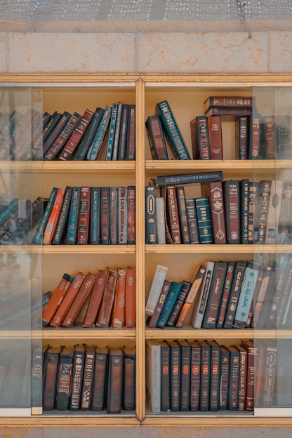 A wooden bookcase filled with various hardcover books, predominantly in shades of red, blue, and brown. The bookshelf has multiple shelves and glass doors, giving the appearance of an organized yet slightly aged collection.
