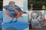 A vibrant street art mural on a brick wall features two large, lifelike bird illustrations. The left side displays a colorful bird with orange and blue plumage, capturing attention with its vivid detail. On the right, a close-up of a bird's face is depicted, showcasing intricate feather details and striking eye expression. The background consists of light blue and white geometric shapes, enhancing the vibrancy of the birds.