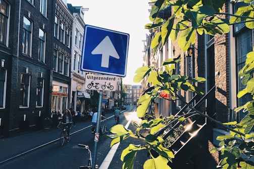 A city street scene with a blue traffic sign displaying an upward arrow and a message in Dutch. Bicycles are present, and people are cycling along the street. Sunlight filters through green leaves from a nearby tree, casting shadows on the brick buildings lining the street.