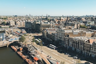 An aerial view of a European city featuring a mix of old and modern architecture. The streets are busy with trams, buses, and cars, while houseboats line a canal. The skyline is dotted with historic buildings and church spires.