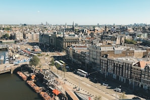 An aerial view of a European city featuring a mix of old and modern architecture. The streets are busy with trams, buses, and cars, while houseboats line a canal. The skyline is dotted with historic buildings and church spires.