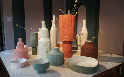 A collection of geometric, paper-like vases and bowls of various sizes displayed on a table. The vases are colored in muted tones such as green, orange, pink, and brown, with some having small yellow ball-like stems protruding from them. A neutral background with vertical green panels enhances the modern aesthetic of the setup.
