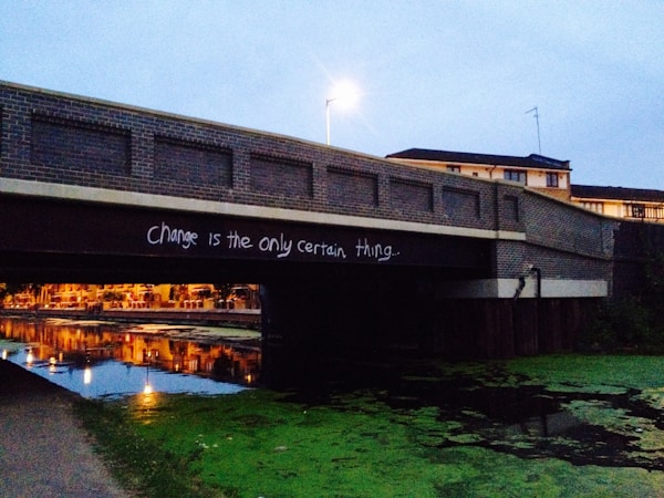 A brick bridge spans over a calm canal with graffiti text stating 'Change is the only certain thing.' The bridge casts a reflection on the water, which is lined with green algae. In the background, a lit streetlamp and buildings are visible against an overcast sky.