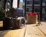 A vintage camera sits on a wooden table, accompanied by a small model of the Eiffel Tower and a round box containing vibrant red roses. In the background, a residential building with balconies can be seen, with sunlight casting shadows.