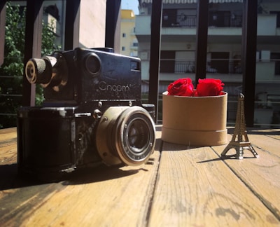 A vintage camera sits on a wooden table, accompanied by a small model of the Eiffel Tower and a round box containing vibrant red roses. In the background, a residential building with balconies can be seen, with sunlight casting shadows.
