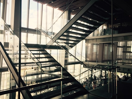 An interior view of a modern building featuring several intersecting glass and metal staircases. The design is sleek and minimalist, with natural light streaming through large windows, creating a pattern of shadows. People can be seen in the background through the glass walls, suggesting a busy urban environment.
