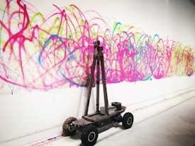 A wall is covered with vibrant graffiti consisting of swirling lines in colors such as pink, blue, yellow, and green. In front of the wall, a small wheeled robot or device is present, possibly used for creating the art.