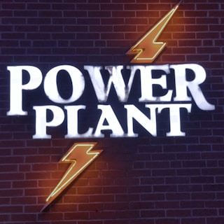 Neon signage on a brick wall displaying the words 'Power Plant' with two bolt symbols, one above and one below the text, illuminated in bright white and orange colors.