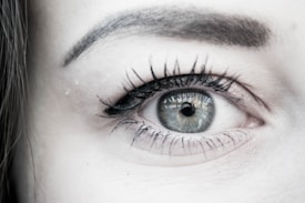 A close-up view of a human eye with detailed focus on the iris, eyelashes, and eyebrow. The iris is grayish in color, and the eyelashes are long and dark. There is a faint reflection visible in the pupil.