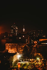 A nighttime cityscape featuring a brightly lit tower in the distance, surrounded by high-rise buildings with illuminated windows. In the foreground, there are residential buildings and a lighted sports court where people can be seen playing.