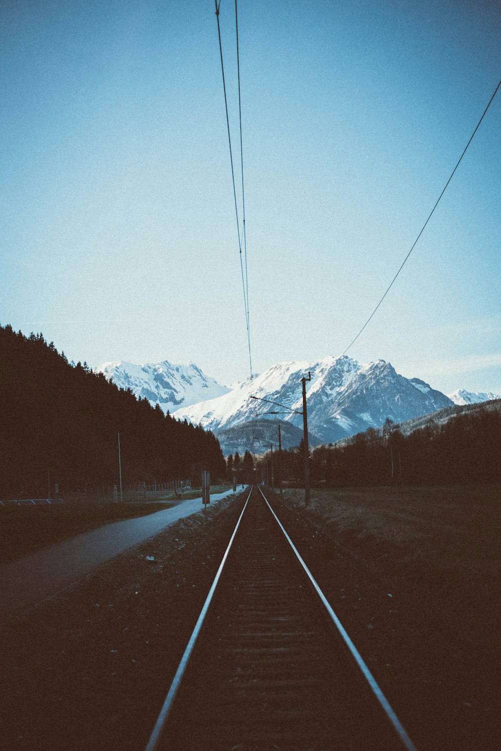 train tracks near road with snow capped mountains background