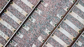 Parallel railway tracks placed on a bed of stones, with wooden sleepers running perpendicular to the tracks. The image shows a diagonal composition, emphasizing the texture and arrangement of the stones and the metallic shine of the tracks.