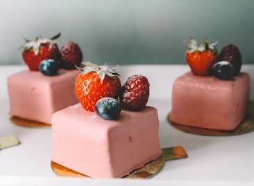 Three square-shaped desserts with pink icing are elegantly decorated with fresh berries, including strawberries, raspberries, and blueberries. The desserts are placed on golden bases and arranged on a white surface.