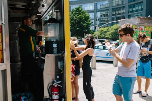 A group of people is gathered around a food truck on a sunny day. The vendor, wearing a green and yellow uniform, is serving food. Customers are holding food items as they stand or walk near the truck. A police car and tall buildings are visible in the background, indicating an urban setting.