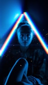 A person is seated in a dark setting with two vibrant, neon light beams forming a triangle around their head. The neon lights emit shades of blue, red, and green, creating a futuristic and edgy atmosphere. The person's face is partially illuminated by the lights, adding a dramatic and moody effect.