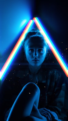 A person is seated in a dark setting with two vibrant, neon light beams forming a triangle around their head. The neon lights emit shades of blue, red, and green, creating a futuristic and edgy atmosphere. The person's face is partially illuminated by the lights, adding a dramatic and moody effect.