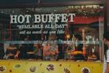 A lively food buffet window display with 'Hot Buffet' and 'Available All Day' written in large letters. Various dishes are visible through the glass, and people inside serve food. The decor includes hanging plants above the window. Reflective surfaces show some street elements outside.