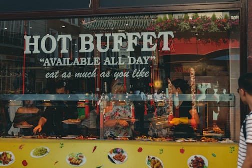 A lively food buffet window display with 'Hot Buffet' and 'Available All Day' written in large letters. Various dishes are visible through the glass, and people inside serve food. The decor includes hanging plants above the window. Reflective surfaces show some street elements outside.
