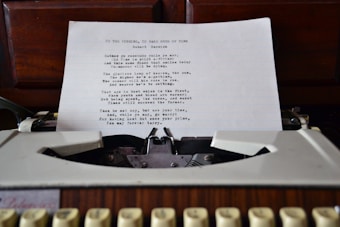 A typewritten sheet of paper is positioned in an old-fashioned typewriter. The sheet contains a poem with the title 'To the Virgins, to Make Much of Time' by Robert Herrick. The typewriter is set against a wooden background likely of a desk or cabinet.