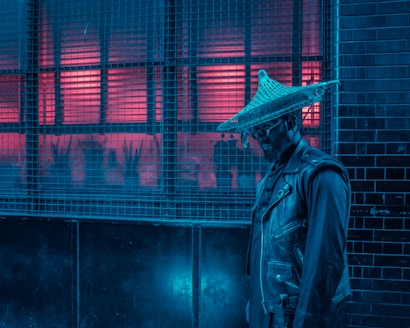 A person wearing a wide-brimmed hat and a leather jacket stands against a gridded wall. The lighting is dramatic with strong blue and red hues, creating a cyberpunk or futuristic feel.