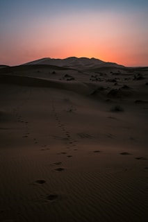 A golden desert dune at sunset with footprints leading into the distance under a glowing orange sky.