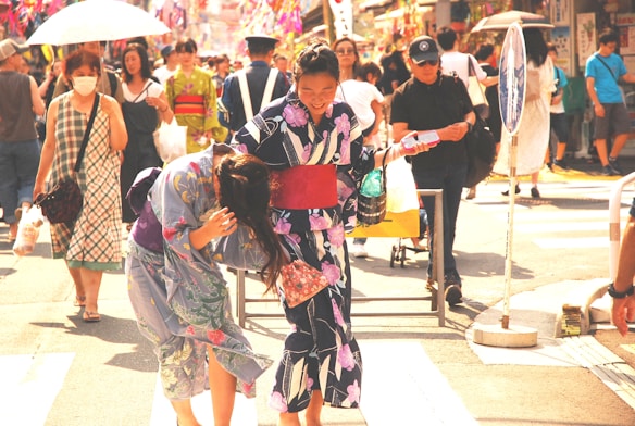 A bustling street scene with people walking. Two women in colorful traditional kimonos are laughing and interacting playfully. One carries a shopping bag and a smaller item while adjusting her kimono. They are surrounded by a diverse crowd, some holding umbrellas and wearing masks, creating a vibrant and lively atmosphere.