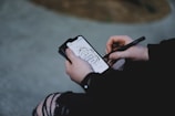 A person is holding a smartphone and using a stylus to draw or write on the screen. The smartphone screen displays an artistic design which includes a motivational quote.