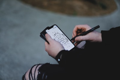 A person is holding a smartphone and using a stylus to draw or write on the screen. The smartphone screen displays an artistic design which includes a motivational quote.