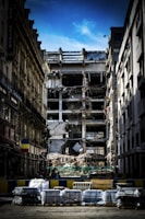 A partially demolished building is surrounded by other intact structures, with debris and construction materials scattered around. The sky is a clear blue, highlighting the contrast between the damaged central building and the neighboring architecture. Barriers and construction equipment are visible, indicating an ongoing demolition or renovation project.