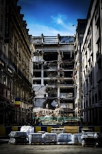 A partially demolished building is surrounded by other intact structures, with debris and construction materials scattered around. The sky is a clear blue, highlighting the contrast between the damaged central building and the neighboring architecture. Barriers and construction equipment are visible, indicating an ongoing demolition or renovation project.