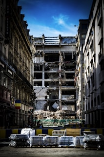 A partially demolished building is surrounded by other intact structures, with debris and construction materials scattered around. The sky is a clear blue, highlighting the contrast between the damaged central building and the neighboring architecture. Barriers and construction equipment are visible, indicating an ongoing demolition or renovation project.