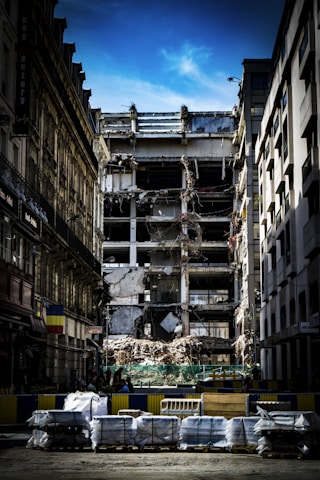 A partially demolished building is surrounded by other intact structures, with debris and construction materials scattered around. The sky is a clear blue, highlighting the contrast between the damaged central building and the neighboring architecture. Barriers and construction equipment are visible, indicating an ongoing demolition or renovation project.