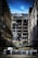 A partially demolished building is surrounded by other intact structures, with debris and construction materials scattered around. The sky is a clear blue, highlighting the contrast between the damaged central building and the neighboring architecture. Barriers and construction equipment are visible, indicating an ongoing demolition or renovation project.