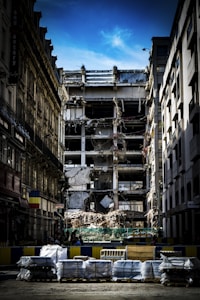 A partially demolished building is surrounded by other intact structures, with debris and construction materials scattered around. The sky is a clear blue, highlighting the contrast between the damaged central building and the neighboring architecture. Barriers and construction equipment are visible, indicating an ongoing demolition or renovation project.