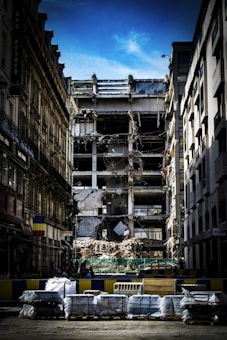 A partially demolished building is surrounded by other intact structures, with debris and construction materials scattered around. The sky is a clear blue, highlighting the contrast between the damaged central building and the neighboring architecture. Barriers and construction equipment are visible, indicating an ongoing demolition or renovation project.